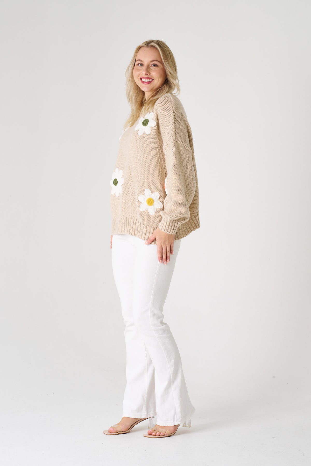 Woman wearing a beige sweater with floral patterns and white pants on a plain background