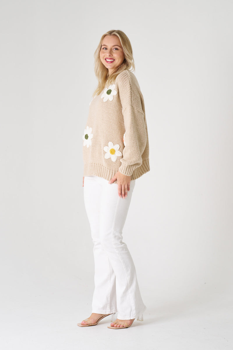 Woman wearing a beige sweater with floral patterns and white pants on a plain background