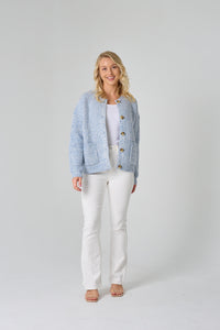 Woman wearing a light blue textured cardigan over a white top and pants on a plain background