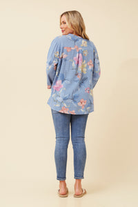 A woman shown from the back wearing a light blue floral button-up shirt and blue skinny jeans, standing against a solid beige studio background.