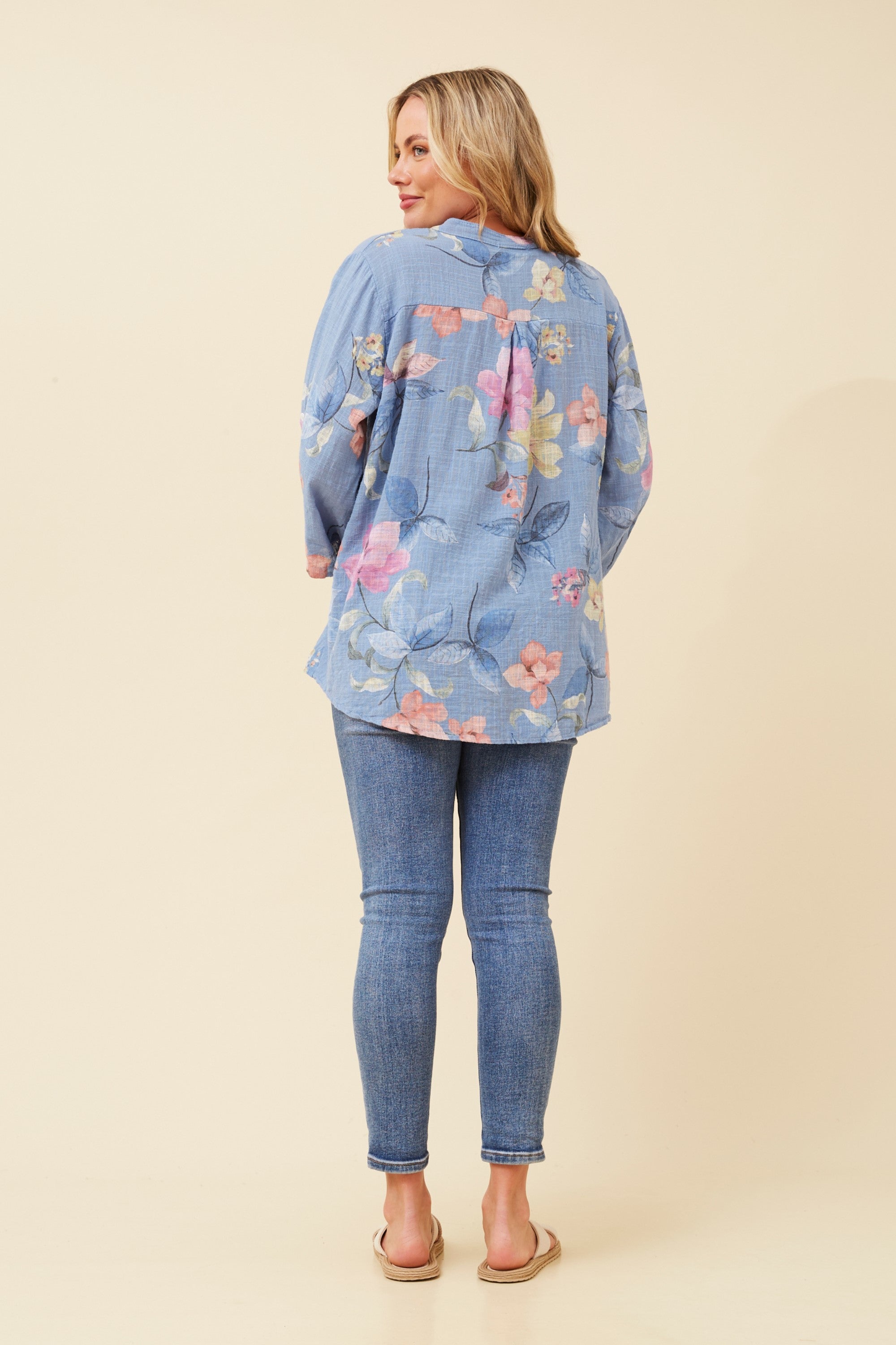 A woman shown from the back wearing a light blue floral button-up shirt and blue skinny jeans, standing against a solid beige studio background.