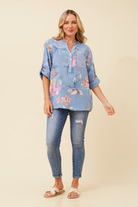 A woman modeling a light blue floral button-up shirt with three-quarter sleeves, paired with blue skinny jeans and white slide sandals, against a solid beige background.