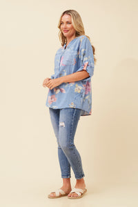 A woman modeling a light blue floral short-sleeve button-up shirt with distressed blue jeans and beige slide sandals, standing against a solid beige studio background.