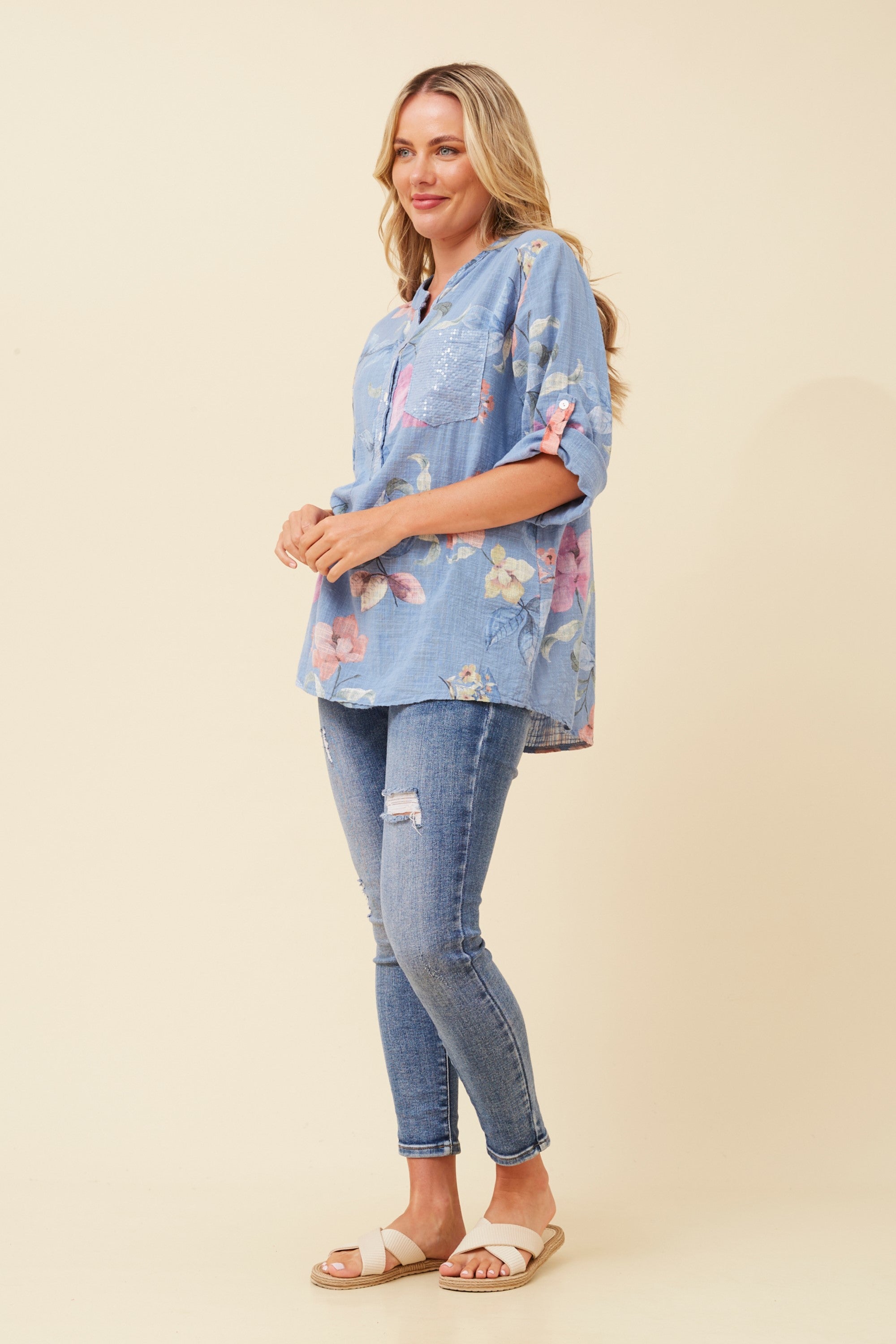 A woman modeling a light blue floral short-sleeve button-up shirt with distressed blue jeans and beige slide sandals, standing against a solid beige studio background.