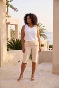 A woman stands outdoors wearing a sleeveless white top and light beige capri pants, with sandals, in a sunlit architectural setting with palm trees and water in the background.