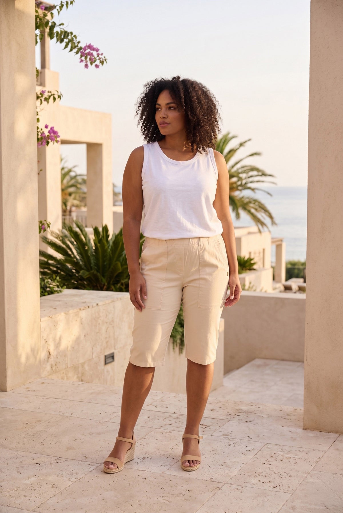 A woman stands outdoors wearing a sleeveless white top and light beige capri pants, with sandals, in a sunlit architectural setting with palm trees and water in the background.