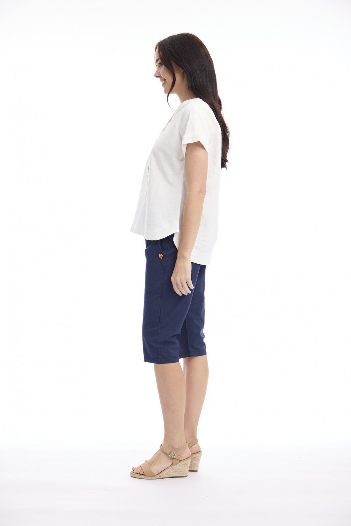 A woman shown in side profile wearing a white short-sleeve top, navy blue knee-length shorts, and beige wedge sandals against a plain white studio background.