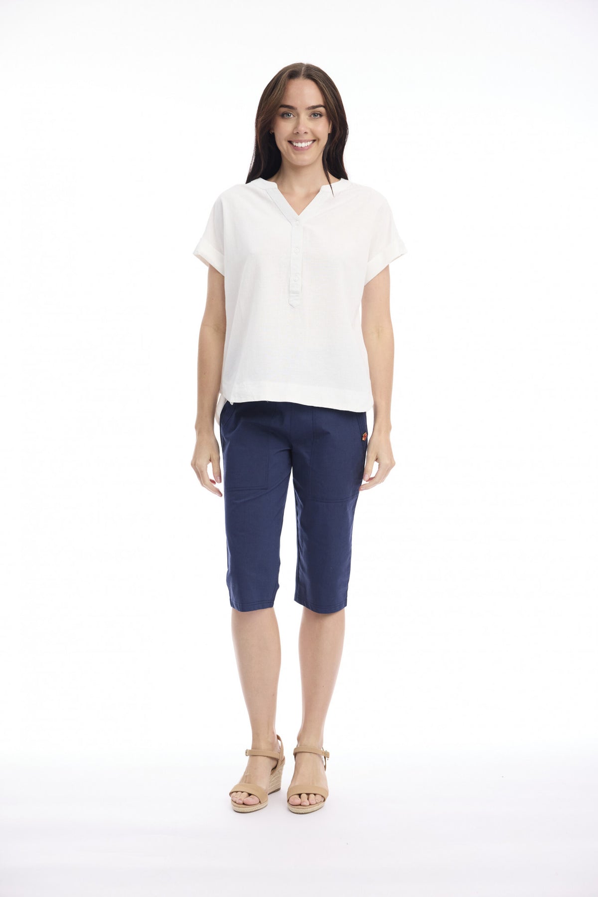 A woman modeling a white short-sleeve collared top and navy blue cropped pants with beige sandals, standing against a plain white studio background.