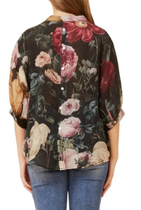 A woman shown from the back wearing a black button-front blouse with a large floral print in pink, red, and beige tones, paired with blue jeans against a plain beige studio background.