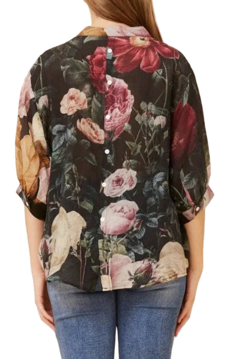 A woman shown from the back wearing a black button-front blouse with a large floral print in pink, red, and beige tones, paired with blue jeans against a plain beige studio background.