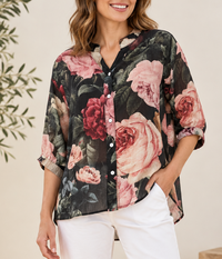 A woman wearing a floral button-up blouse with pink and red roses on a dark background, paired with white pants, photographed in a softly lit indoor setting.