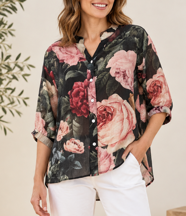 A woman wearing a floral button-up blouse with pink and red roses on a dark background, paired with white pants, photographed in a softly lit indoor setting.