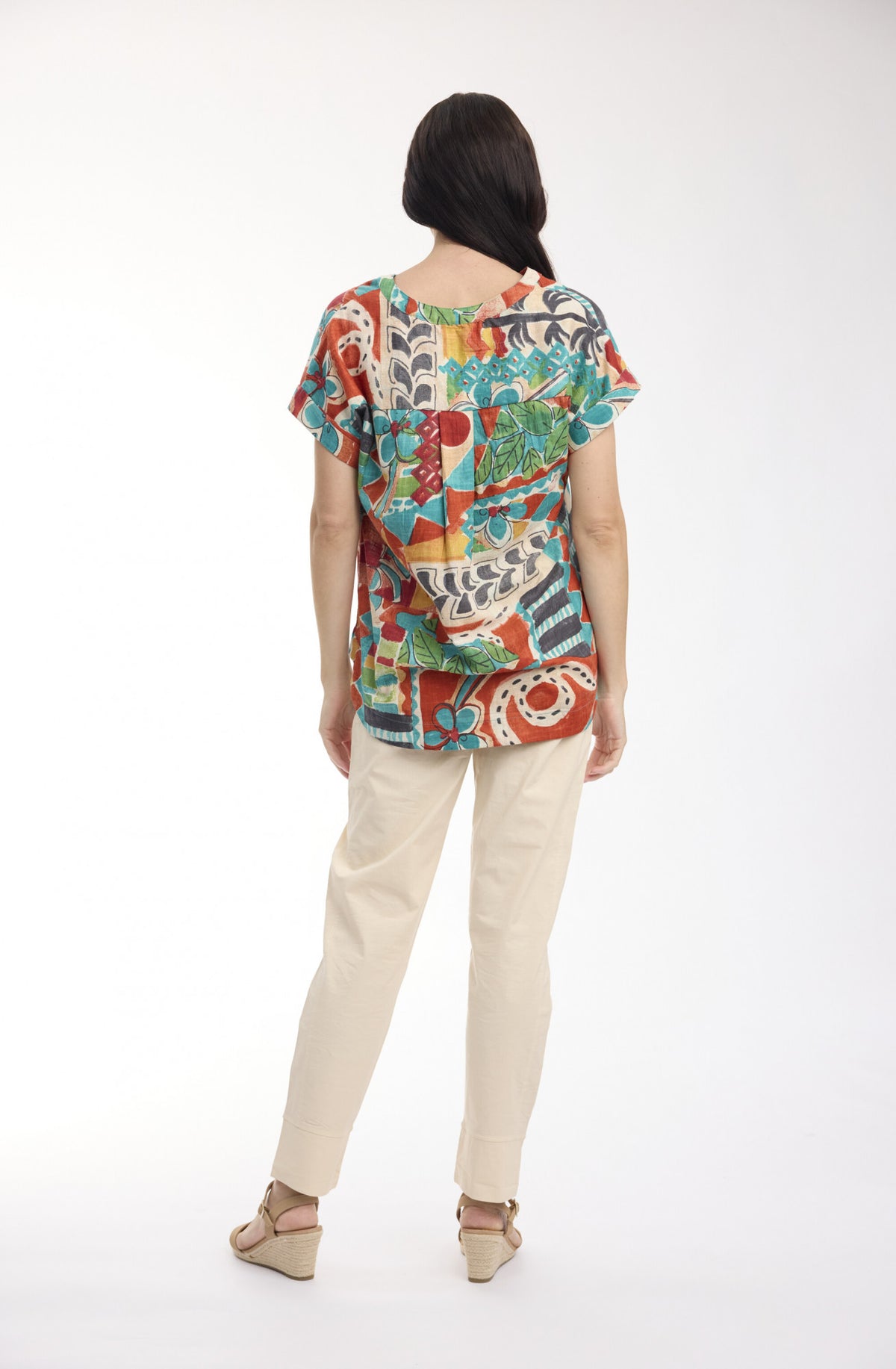 A woman viewed from the back wearing a colorful short-sleeve top with an abstract multicolor print, paired with light beige pants and beige wedge sandals, against a plain white studio background.