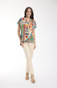 A woman modeling a short-sleeve button-up shirt with a vibrant multicolor tropical and abstract print, paired with beige pants and tan sandals, against a plain white studio background.