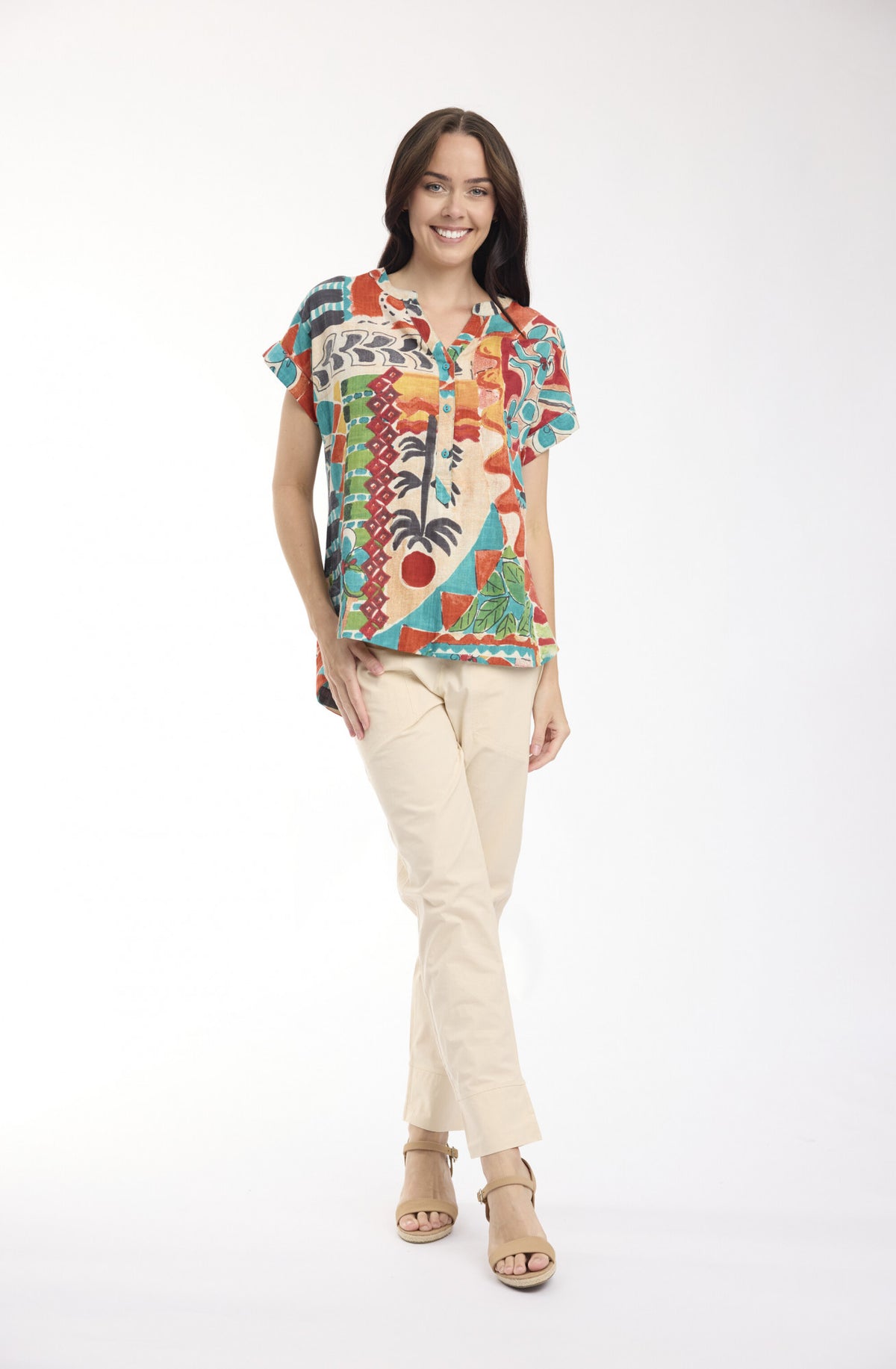 A woman modeling a short-sleeve button-up shirt with a vibrant multicolor tropical and abstract print, paired with beige pants and tan sandals, against a plain white studio background.