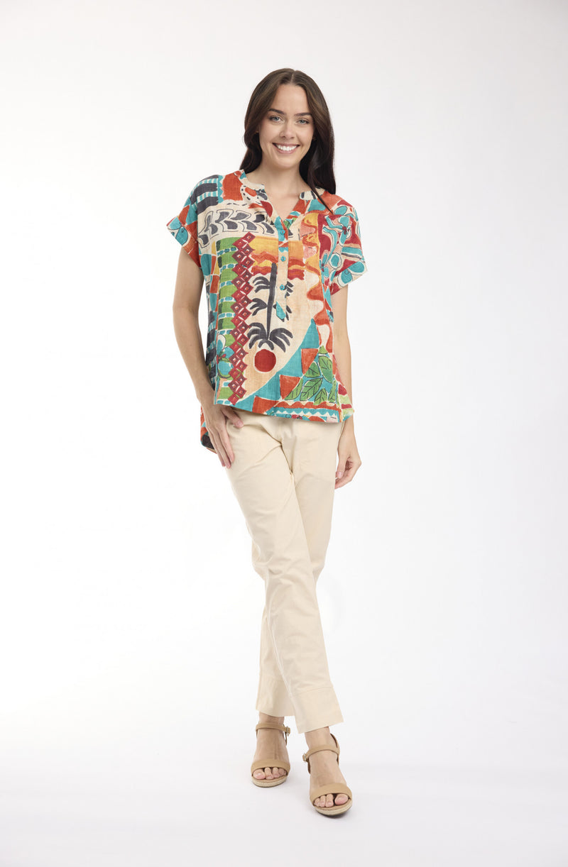 A woman modeling a short-sleeve button-up shirt with a vibrant multicolor tropical and abstract print, paired with beige pants and tan sandals, against a plain white studio background.