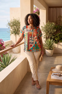 A woman stands on a sunlit balcony overlooking the ocean, wearing a colorful short-sleeve button-front top with a bold tropical and floral print, cream pants, and tan sandals. She holds a small beige clutch, with potted plants, a wooden table, and a hat nearby.