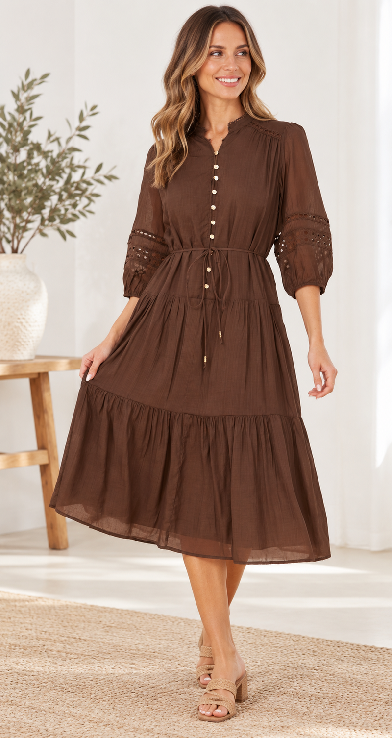 A woman modeling a brown midi dress with a tiered skirt, buttoned front, and lace-up waist tie in a bright indoor setting.